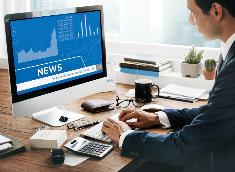 Man in a suit typing on a keyboard at a desk with a computer screen displaying news and financial charts.