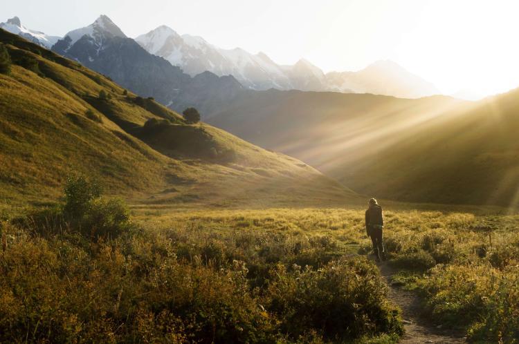 A person walks through a sunlit grassy valley with rolling hills and distant mountains, as golden sunlight streams across the landscape at sunrise or sunset.