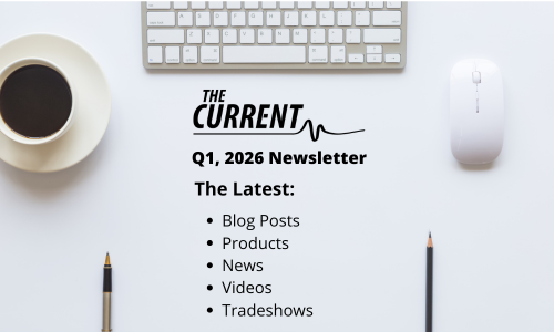 Top-down view of a white desk with coffee, keyboard, mouse, and pens. Text: "THE CURRENT Q1, 2026 Newsletter. The Latest: Blog Posts, Products, News, Videos, Tradeshows."