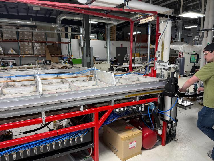 A man operates an industrial lamination machine. Boxes and tools are visible underneath.